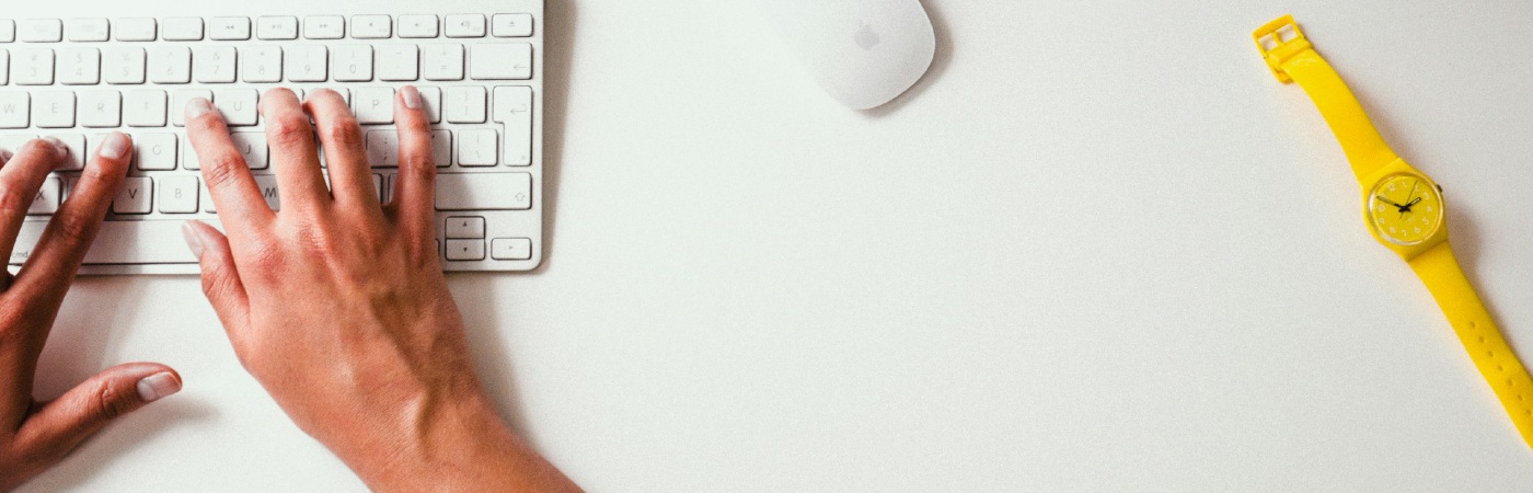Hands typing on a white keyboard next to a white Apple Magic Mouse, with a yellow wristwatch and smartphone on a clean white desk.
